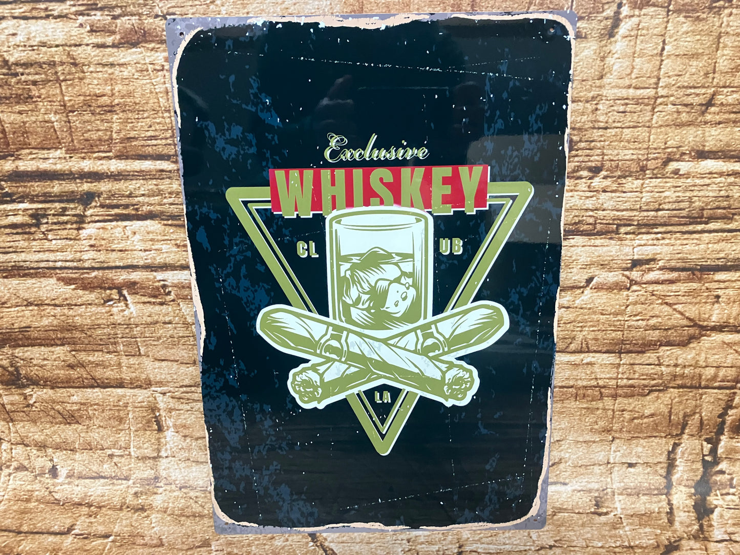 Vintage metalen bord met een zwarte achtergrond en verweerde randen, waarop een glas whisky met ijs staat afgebeeld binnen een driehoekig frame. Voor het glas liggen twee gekruiste sigaren. Boven en onder de illustratie staat de tekst “Exclusive Whiskey Club LA”, waarbij het woord “Whiskey” groot en opvallend in rood‑witte letters is vormgegeven. Het geheel hangt tegen een houten achtergrond en straalt een luxe, klassieke bar‑ of lounge‑sfeer uit.