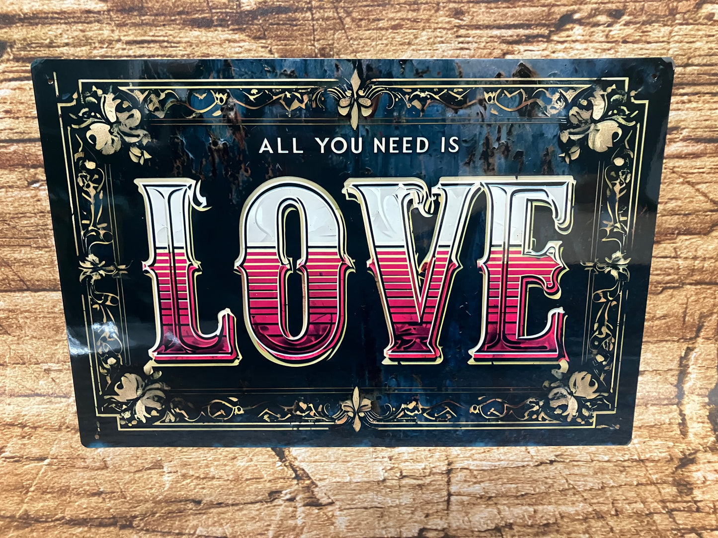 Decoratief bord met de tekst “ALL YOU NEED IS LOVE” in witte en roodgestreepte letters, omlijst met sierlijke gouden randen op een donkere achtergrond, gemonteerd op hout voor een warme en nostalgische uitstraling.