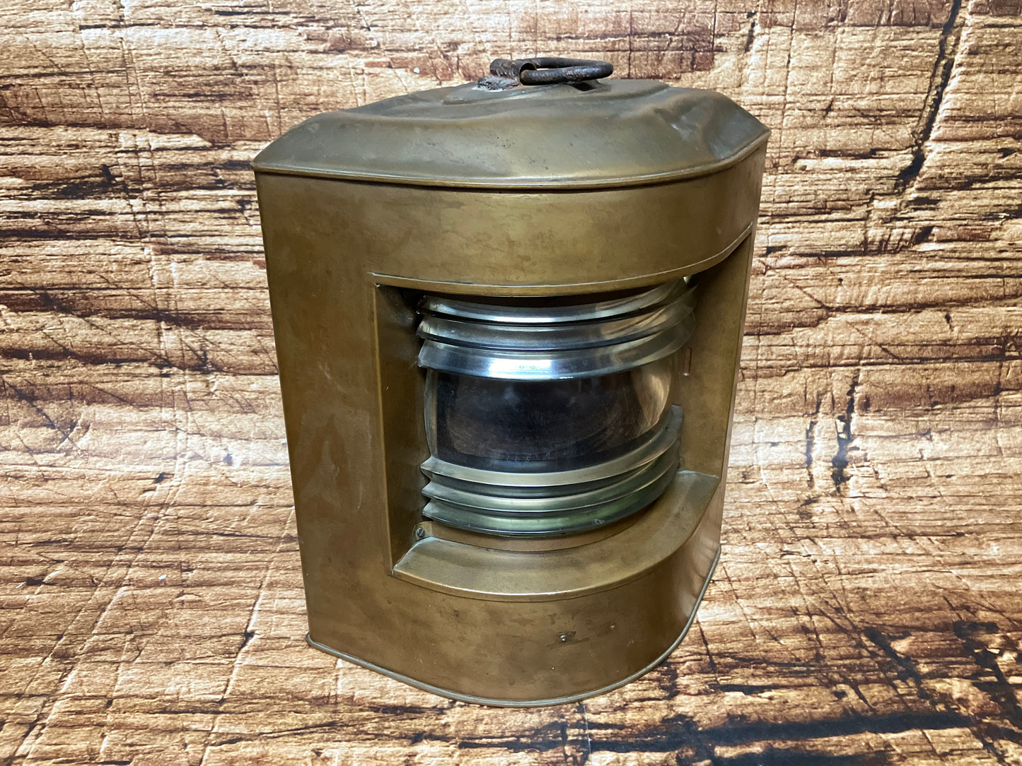 “Antieke messing bakboordlamp met gebogen glazen lens en karakteristieke patina, ontworpen voor scheepsnavigatie, staand op een lichte houten ondergrond. De robuuste constructie en historische details benadrukken het authentieke maritieme karakter van het object.”