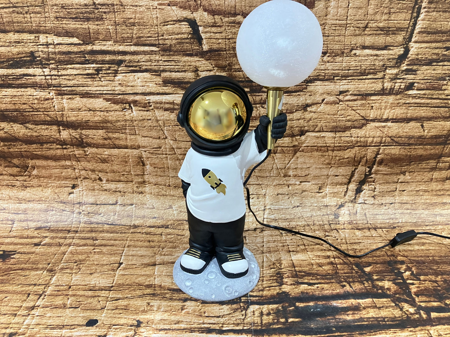 Een decoratieve astronautlamp met een goudkleurige vizier, staand op een maanachtig voetstuk, terwijl de astronaut een ronde, gloeiende bolvormige lamp omhoog houdt