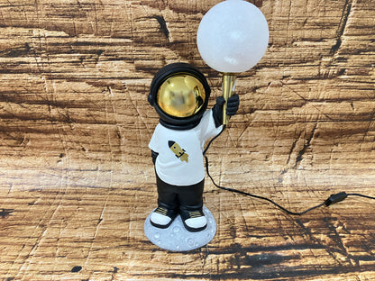 Een decoratieve astronautlamp met een goudkleurige vizier, staand op een maanachtig voetstuk, terwijl de astronaut een ronde, gloeiende bolvormige lamp omhoog houdt