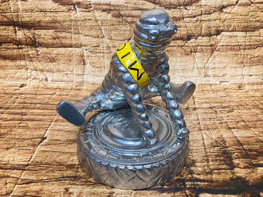 Metalen figurine van het Michelin-mannetje (Bibendum) zittend op een gedetailleerde autoband, met een zilveren afwerking en een gele sjerp met de letters "MICHELIN";.