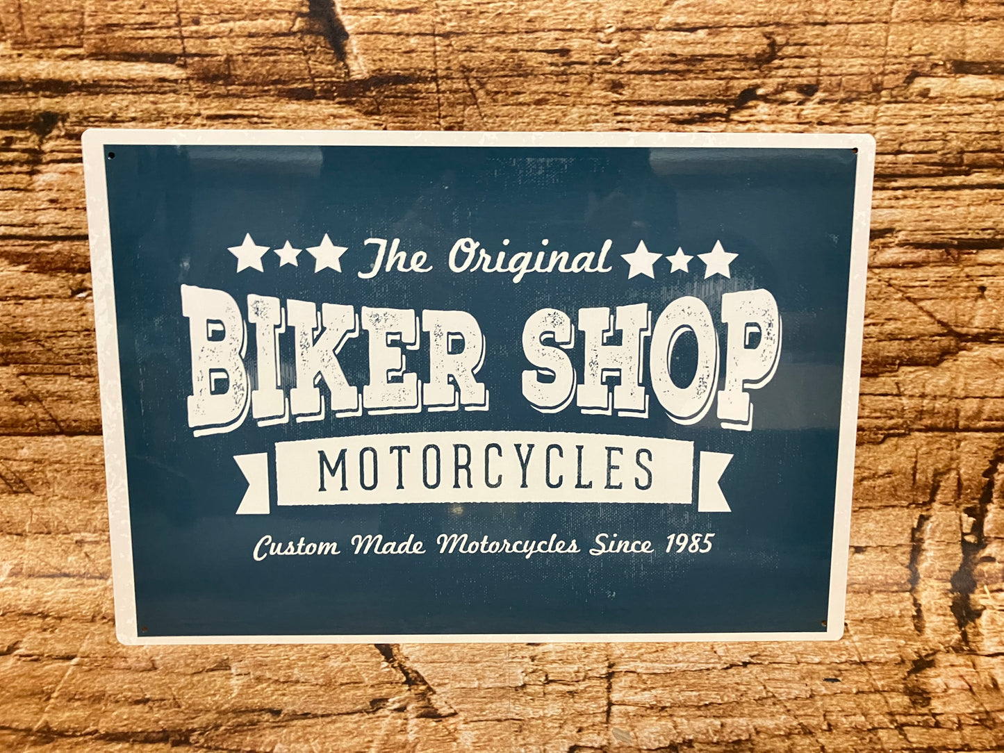 Vintage metalen bord met donkerblauwe achtergrond en witte tekst “★ The Original ★ BIKER SHOP – MOTORCYCLES – Custom Made Motorcycles Since 1985”, omlijst met decoratieve elementen en gemonteerd op een houten oppervlak.