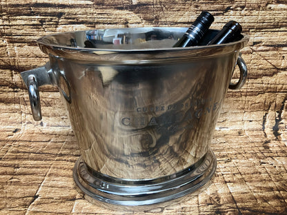 Een glanzende metalen champagne‑emmer met twee flessen erin staat op een houten ondergrond. De emmer heeft twee stevige handgrepen en een elegante gravure met de tekst “Cuvée de Prestige – Champagne”. De reflecterende metalen afwerking en de warme houtstructuur op de achtergrond geven het geheel een luxe en feestelijke uitstraling.