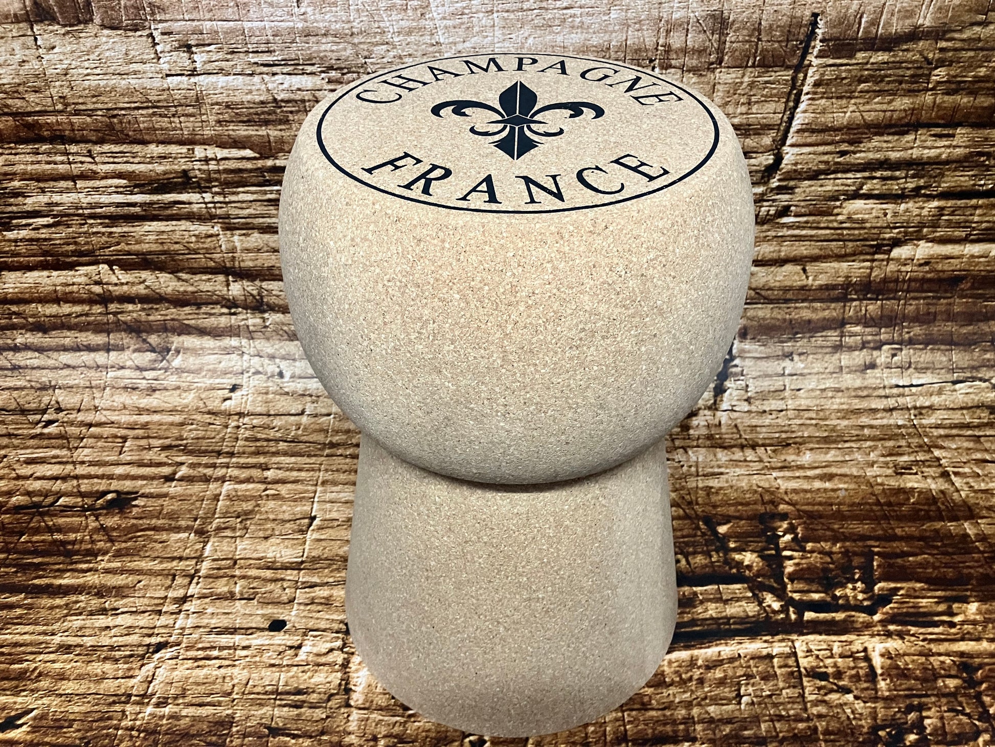 Kruk in de vorm van een grote champagnekurk met het stempel “Champagne France”, geplaatst op een rustieke houten ondergrond; uniek designmeubel voor interieur, wijnbar of horeca.