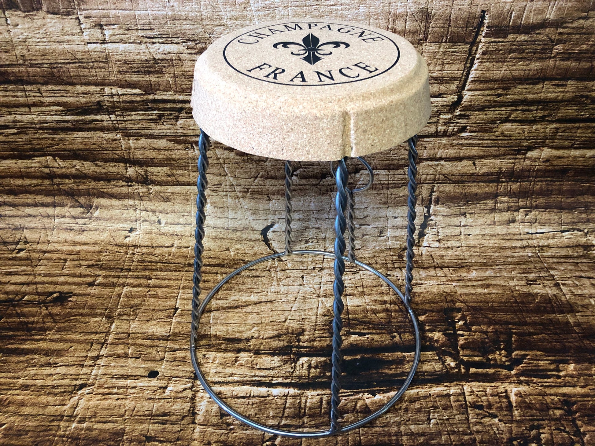 Kruk in de vorm van een champagnekurk met metalen draadpoten die het muselet‑frame van een champagnefles nabootsen, met op de zitting de opdruk “Champagne France”, geplaatst op een rustieke houten ondergrond.
