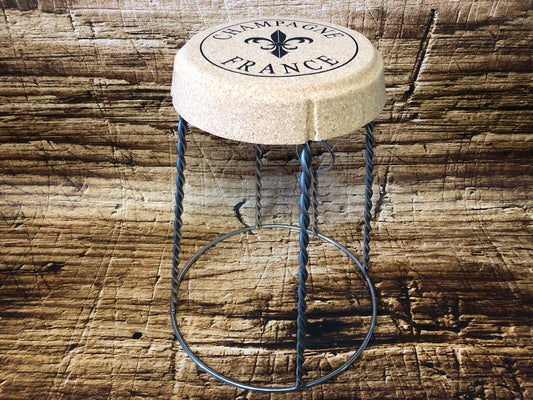 Kruk in de vorm van een champagnekurk met metalen draadpoten die het muselet‑frame van een champagnefles nabootsen, met op de zitting de opdruk “Champagne France”, geplaatst op een rustieke houten ondergrond.