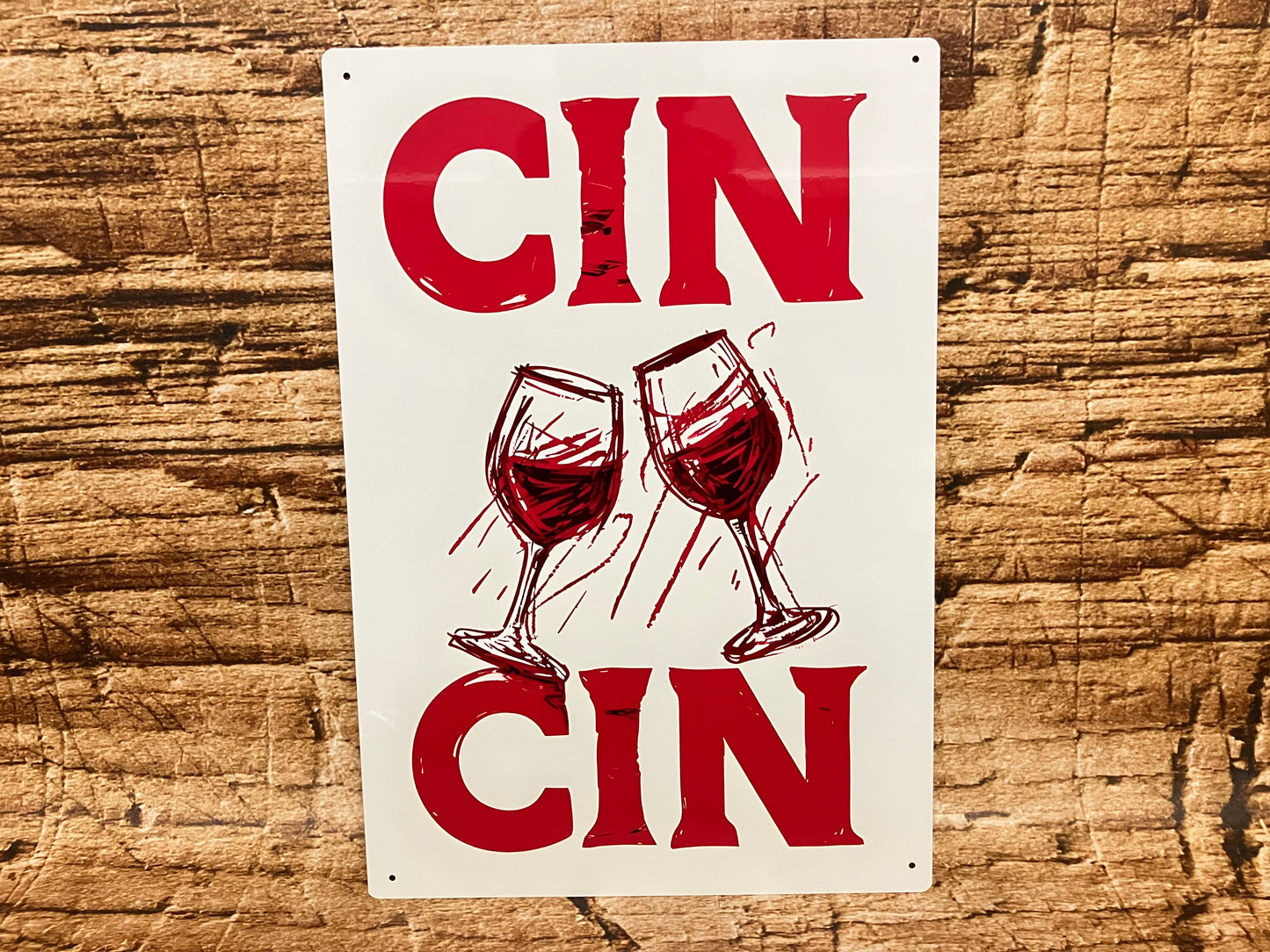 Decoratief bord met de tekst “CIN CIN” in rode letters boven en onder een illustratie van twee proostende wijnglazen, op een rustieke houten achtergrond met feestelijke uitstraling.