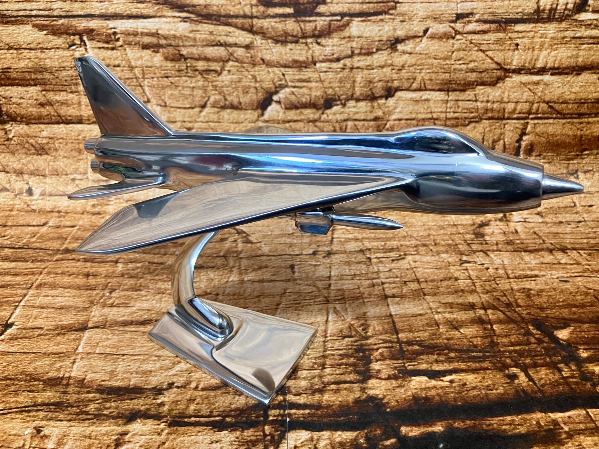 Glanzend metalen model van een straaljager met strakke, hoekige lijnen, gemonteerd op een gebogen standaard met een vierkante voet. Het vliegtuig heeft een gestroomlijnd, futuristisch silhouet en staat tegen een rustieke houten achtergrond, waardoor het gepolijste metaal extra opvalt. De combinatie van het strakke ontwerp en de warme textuur van de ondergrond geeft het geheel een luxe, decoratieve uitstraling.