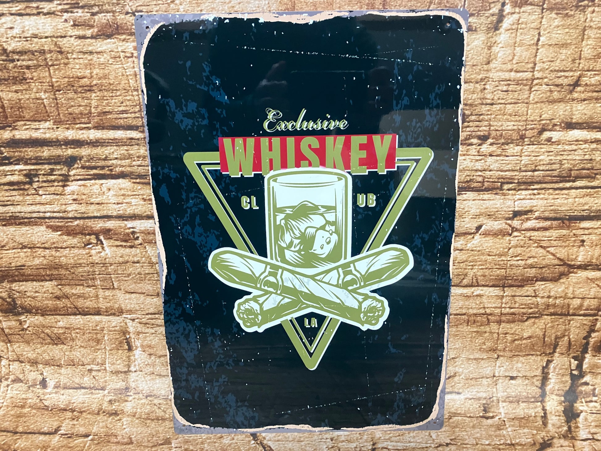 Vintage metalen bord met een zwarte achtergrond en verweerde randen, waarop een glas whisky met ijs staat afgebeeld binnen een driehoekig frame. Voor het glas liggen twee gekruiste sigaren. Boven en onder de illustratie staat de tekst “Exclusive Whiskey Club LA”, waarbij het woord “Whiskey” groot en opvallend in rood‑witte letters is vormgegeven. Het geheel hangt tegen een houten achtergrond en straalt een luxe, klassieke bar‑ of lounge‑sfeer uit.
