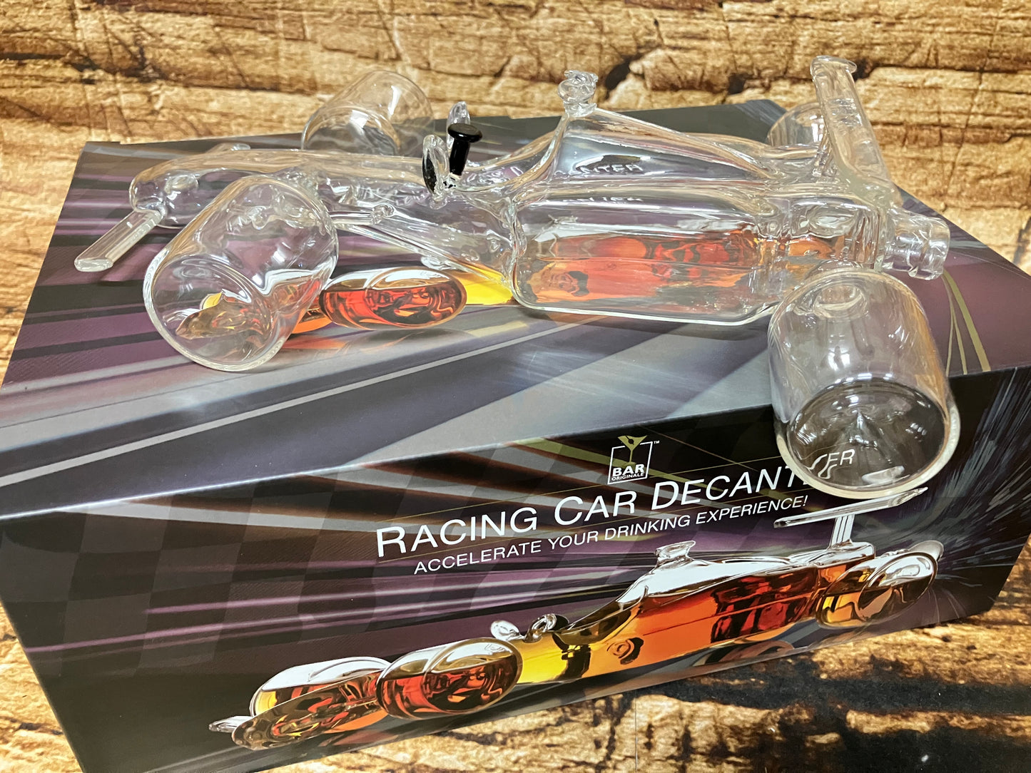 "Een glazen decanter in de vorm van een raceauto staat boven op de bijbehorende verpakking. De decanter is volledig transparant en heeft vier afzonderlijke glazen wielen en een stopper bovenop, waar normaal de cockpit zou zitten. De doos toont een afbeelding van dezelfde raceauto‑decanter, maar dan gevuld met een amberkleurige drank, waardoor de vormen en details extra opvallen.