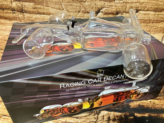 "Een glazen decanter in de vorm van een raceauto staat boven op de bijbehorende verpakking. De decanter is volledig transparant en heeft vier afzonderlijke glazen wielen en een stopper bovenop, waar normaal de cockpit zou zitten. De doos toont een afbeelding van dezelfde raceauto‑decanter, maar dan gevuld met een amberkleurige drank, waardoor de vormen en details extra opvallen.