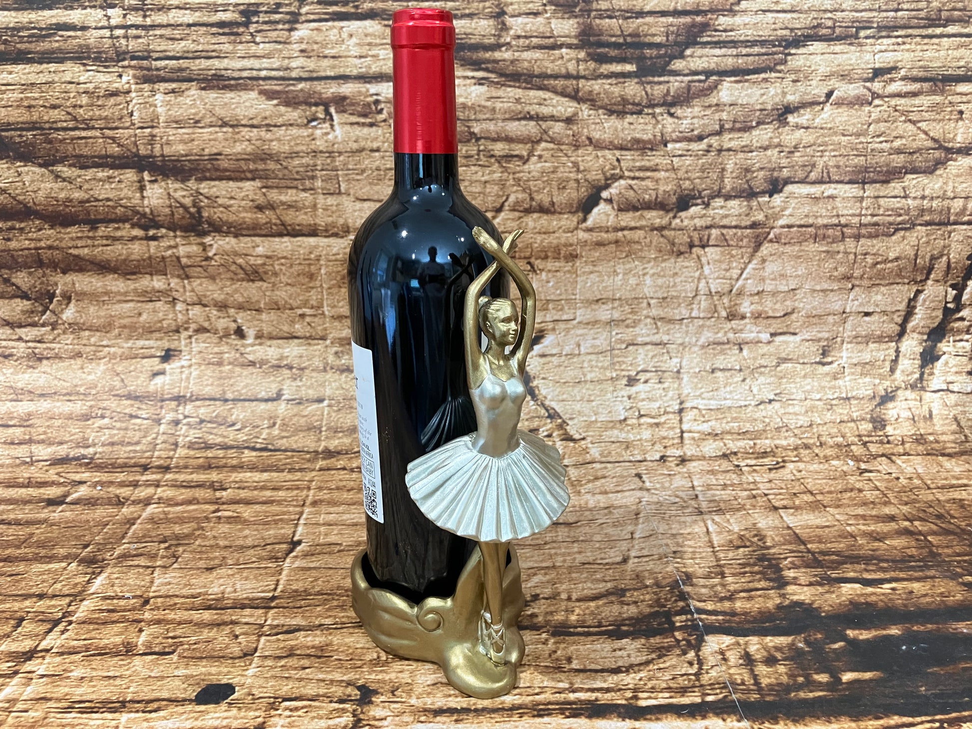 Alt‑tekst:
Een gouden ballerina wijnhouder in elegante pointe‑pose ondersteunt een donkere wijnfles met rode capsule