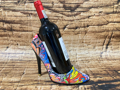 Alt‑tekst:
Een decoratieve wijnhouder in de vorm van een hoge hak met een kleurrijk graffiti‑design, waarin een donkere wijnfles met rode capsule rechtop staat