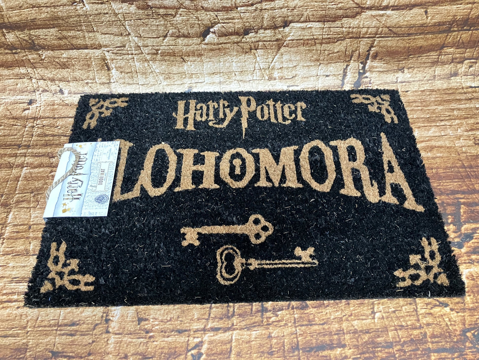 Zwarte Harry Potter‑deurmat met het woord “Alohomora” in goudkleurige letters, twee vintage sleutels en decoratieve hoeken.