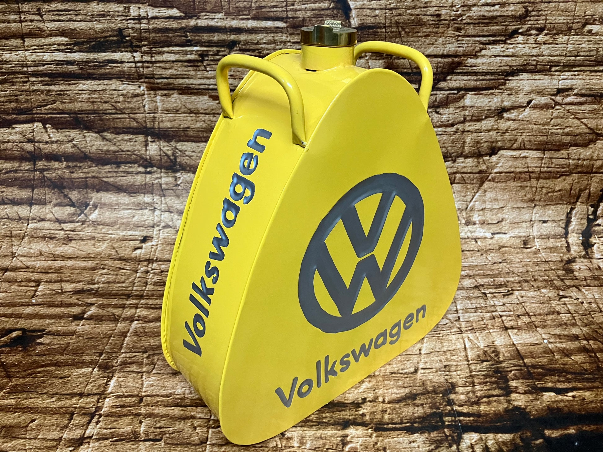 "Gele vintage Volkswagen metalen brandstof- of oliekannister met grijs VW‑logo, driehoekige vorm, messing dop en dubbele handgrepen, gebruikt als oldtimer- en garage‑decoratie."
