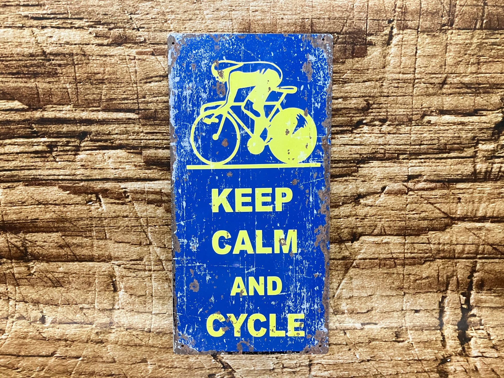  Vintage metalen bord met blauwe achtergrond, gele tekst “KEEP CALM AND CYCLE” en een gestileerde fietser bovenaan, gemonteerd op een houten oppervlak met verweerde uitstraling.