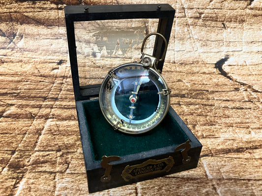 "Vintage metalen kompas in houten box met glasplaat, groene viltvoering en label ‘Spencer & Co. London 1905’, gebruikt als decoratief en verzamelobject."