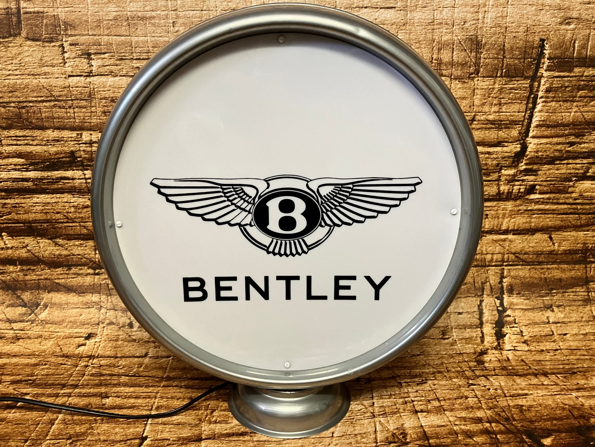 Een ronde, verlichte Bentley-lichtbak met een zilverkleurige metalen rand, gefotografeerd tegen een achtergrond van horizontaal gestapelde houten planken. In het midden van het witte, opaalachtige glaspaneel prijkt het iconische zwarte 'Winged B'-logo van Bentley: een hoofdletter 'B' omsloten door een krans en geflankeerd door twee gestileerde vlerken. Direct daaronder staat de merknaam 'BENTLEY' in een strak, zwart schreefloos lettertype. De lichtbak rust op een kleine, ronde metalen voet