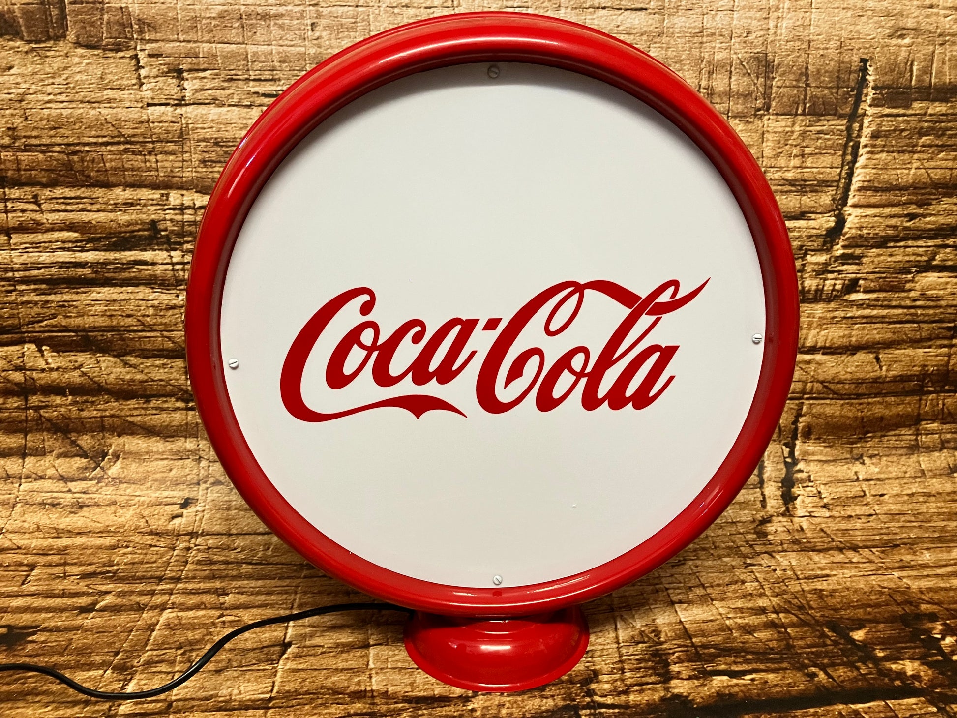 Een ronde, verlichte Coca-Cola lichtreclame in de stijl van een klassieke benzinepomp-globe. Het object heeft een dieprood metalen frame en een bolvormig, wit doorschijnend paneel in het midden. Op het witte paneel staat het iconische Coca-Cola logo in het rood, uitgevoerd in het kenmerkende sierlijke cursieve lettertype. De lichtbak rust op een bijbehorende rode, ronde voet en straalt een egale, warme witte verlichting uit van binnenuit.