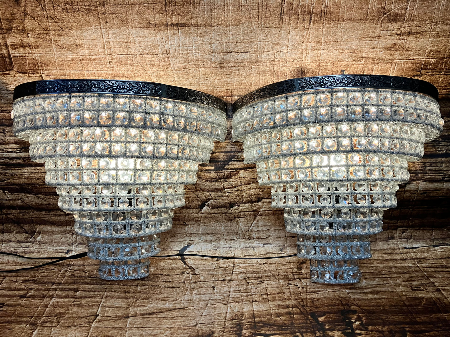 “Twee luxe kristallen wandlampen met donkere metalen rand en warm licht, gemonteerd tegen een houten achtergrond.”