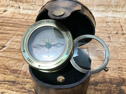   
"Een vintage messing kompas uit 1941, vervaardigd door Henry Hughes Ltd. in Londen, ligt open in een ronde leren etui op een houten ondergrond. De kompasroos toont duidelijke windrichtingen en gradenmarkeringen, terwijl een fijn naaldje naar het noorden wijst. De leren behuizing heeft een knoopsluiting en een kleine ingebouwde vergrootlens die boven het glas rust, wat het instrument een authentieke, historische uitstraling geeft. De warme tinten van het messing en het verweerde leer benadrukken het antie