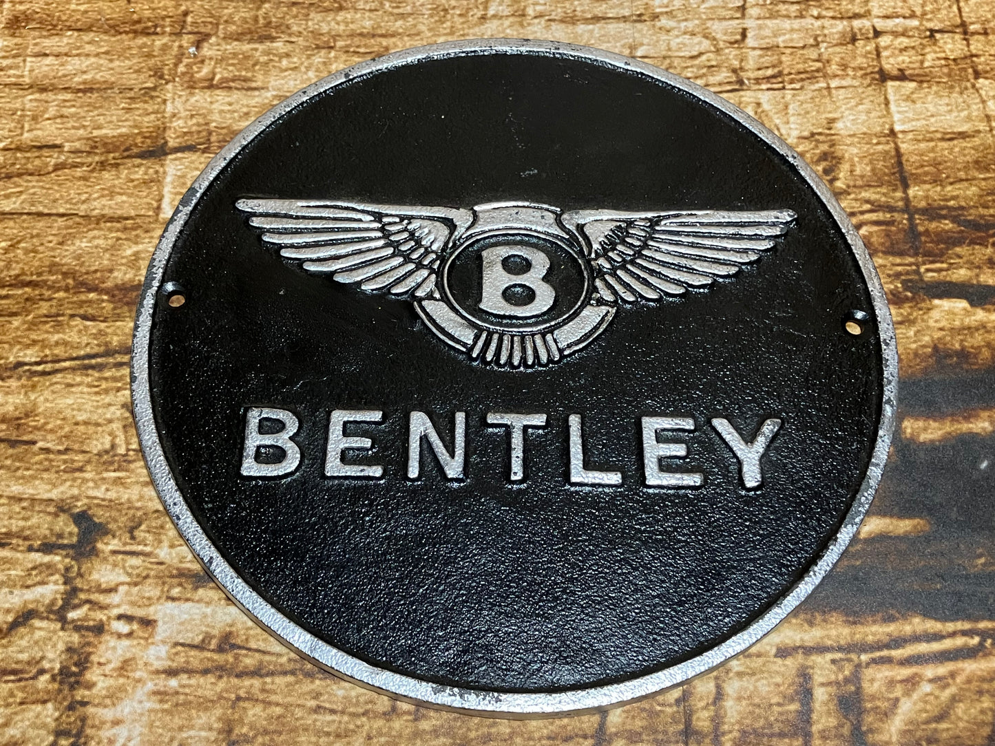 Ronde metalen wandplaat met het zilveren Bentley‑logo op zwarte achtergrond, geschikt als luxe automotive wanddecoratie.