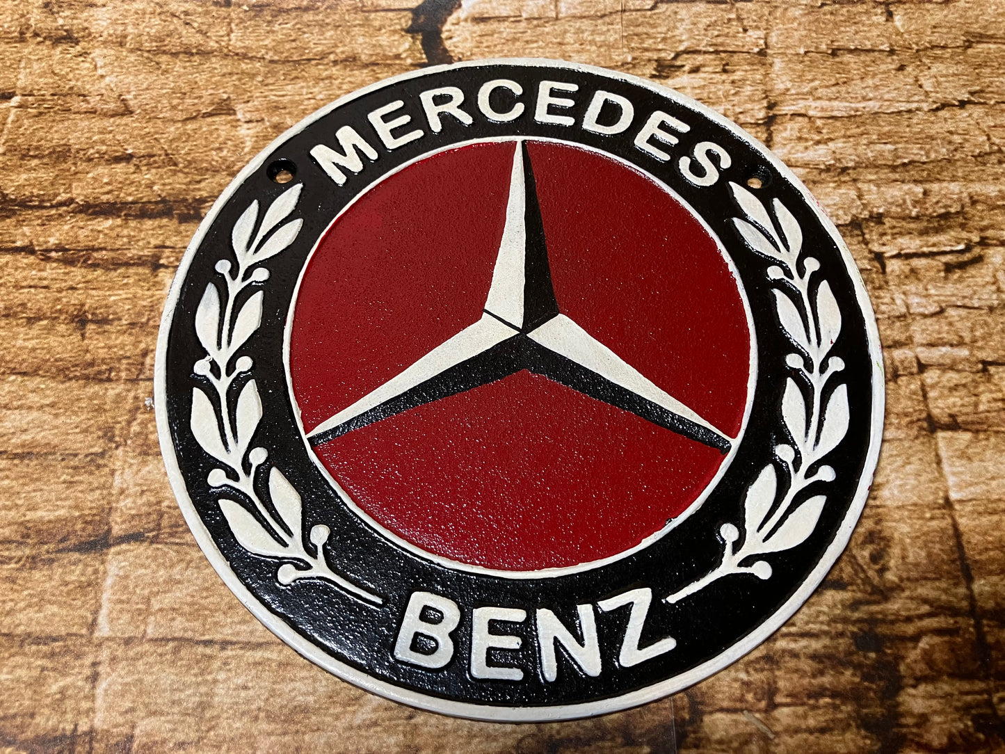 Metalen Mercedes‑Benz embleem met rode achtergrond, zilveren ster en zwarte rand met laurierbladeren.