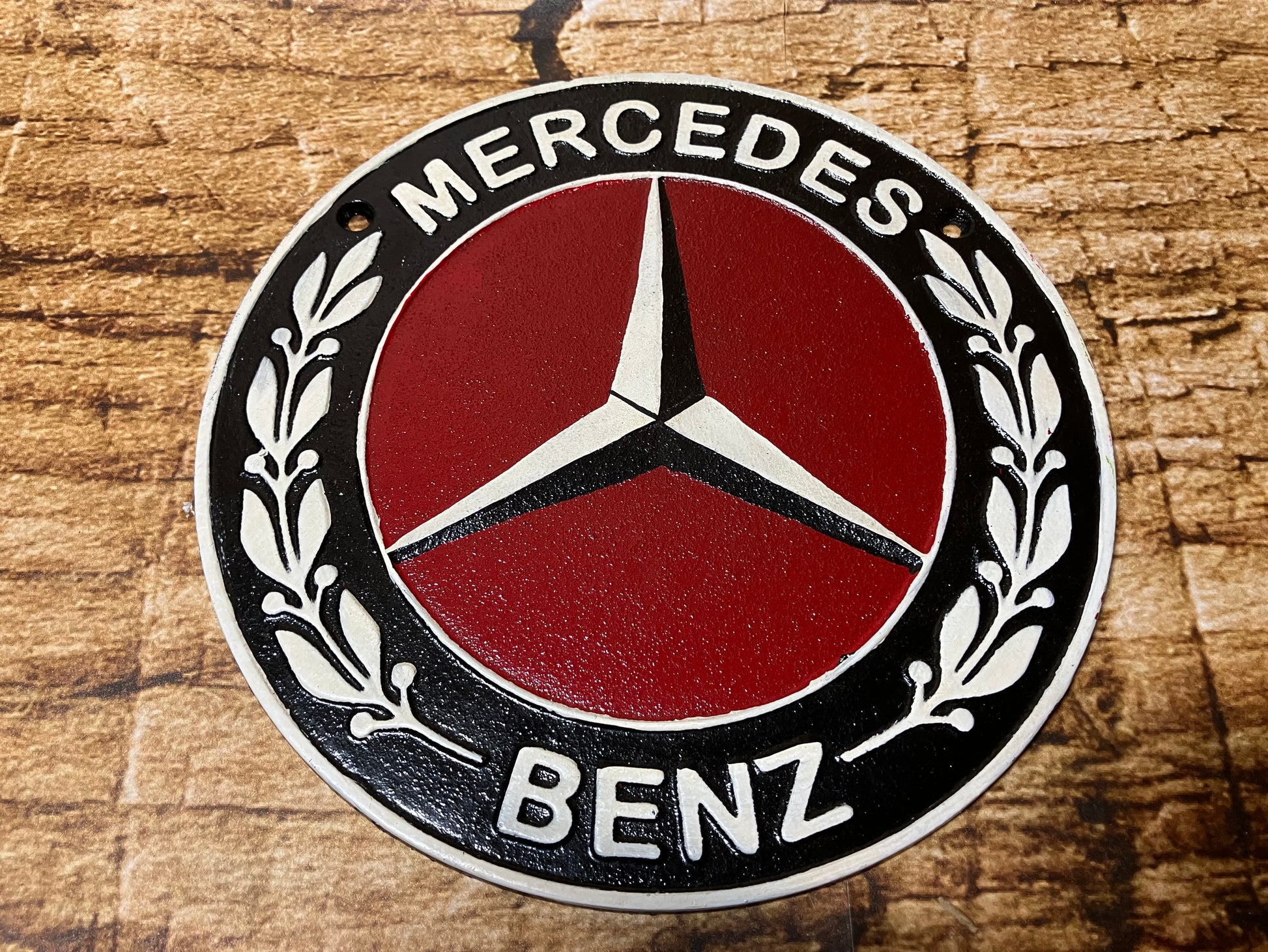 Metalen Mercedes‑Benz embleem met rode achtergrond, zilveren ster en zwarte rand met laurierbladeren.