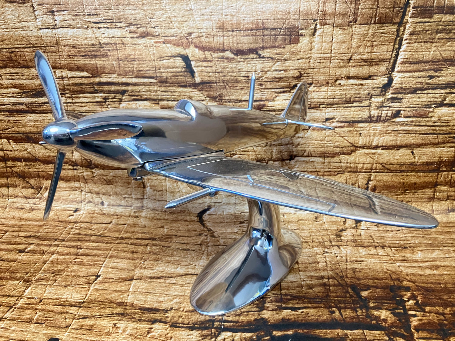 "Aluminium vliegtuigmodel op een gebogen standaard, gefotografeerd op een houten ondergrond; decoratief object met vintage jachtvliegtuig‑design."