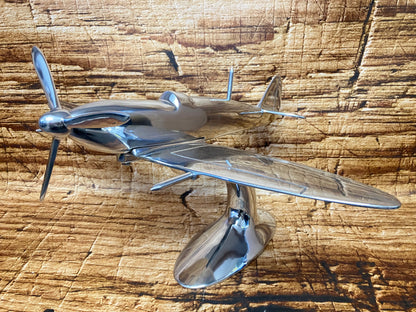 "Aluminium vliegtuigmodel op een gebogen standaard, gefotografeerd op een houten ondergrond; decoratief object met vintage jachtvliegtuig‑design."