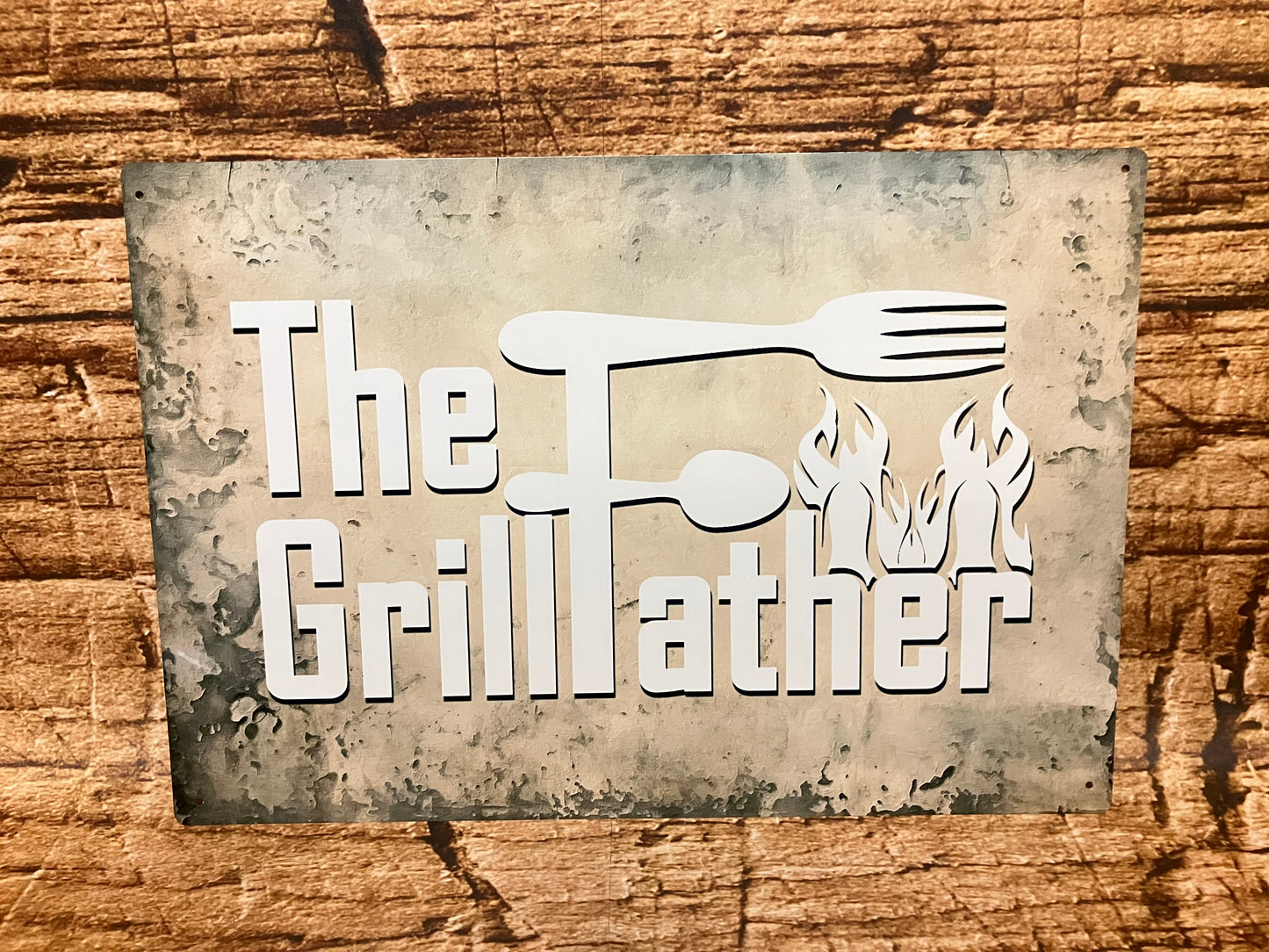 Decoratief bord met de tekst “The GrillFather” in witte letters op een donkere, verweerde achtergrond, met een vork en lepel verwerkt in de letter F en vlammen aan de onderzijde, als humoristische knipoog naar barbecuefans.