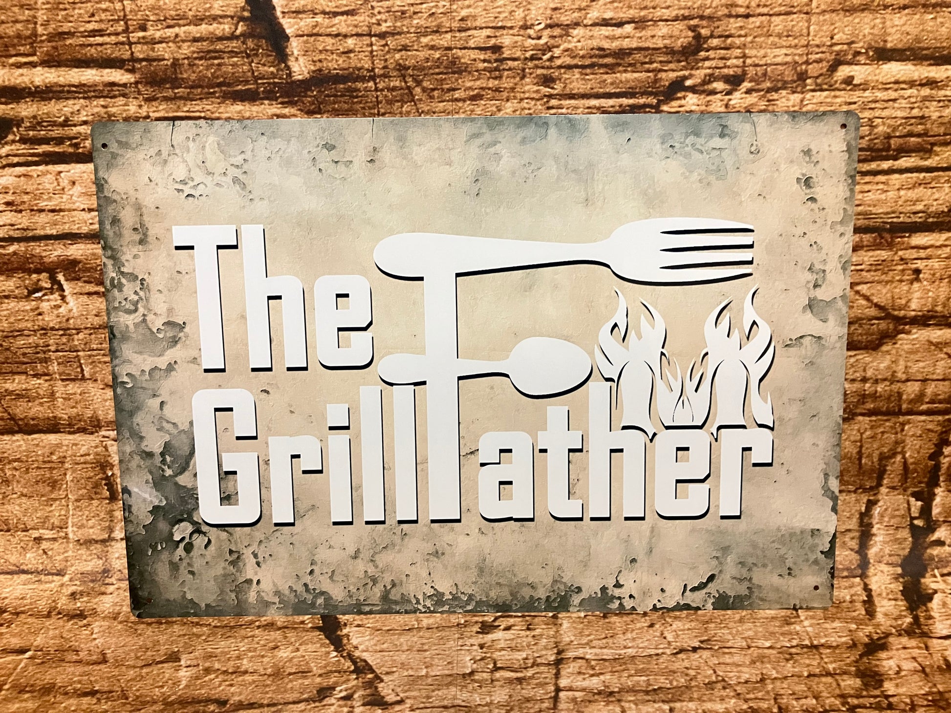 Decoratief bord met de tekst “The GrillFather” in witte letters op een donkere, verweerde achtergrond, met een vork en lepel verwerkt in de letter F en vlammen aan de onderzijde, als humoristische knipoog naar barbecuefans.