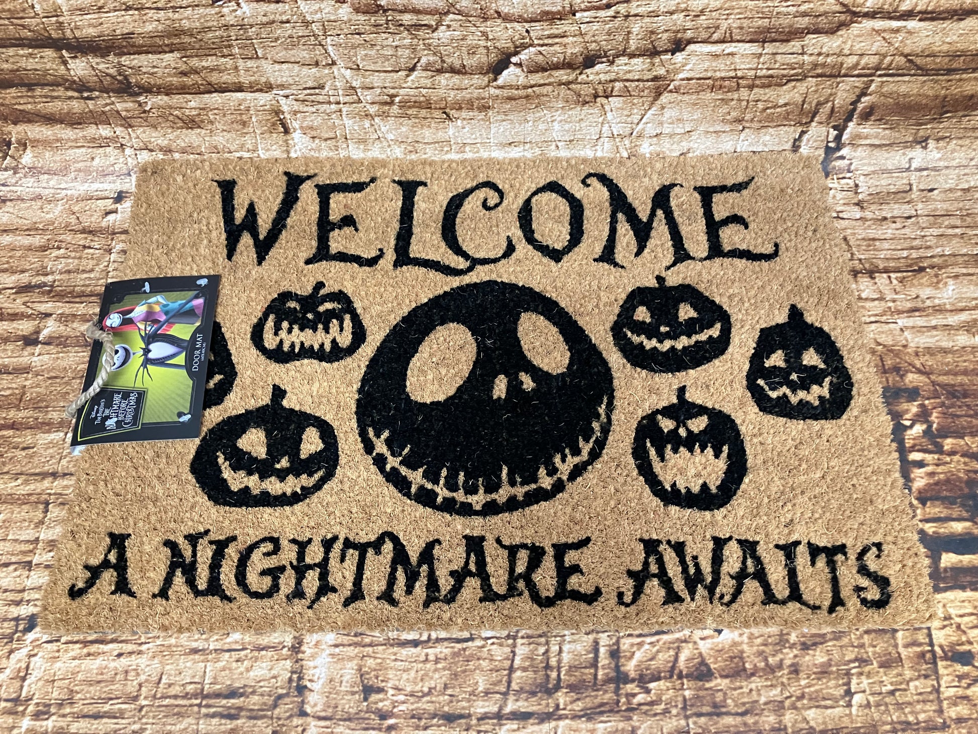 Beige Halloween‑deurmat met groot Jack‑gezicht en de tekst “Welcome – A Nightmare Awaits”, omringd door kleine pompoengezichten.