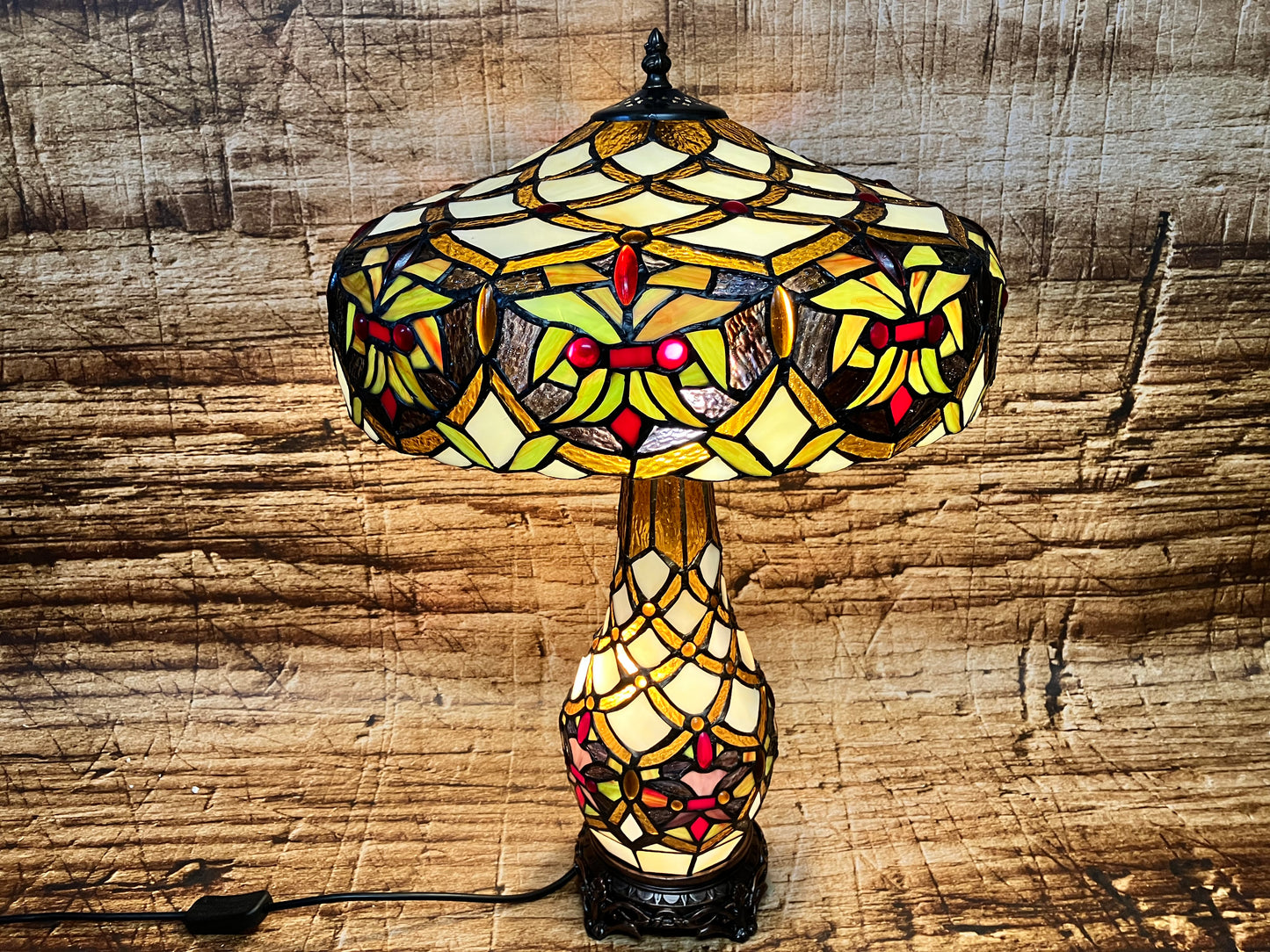 Tiffany-stijl tafellamp met kleurrijke glazen kap in geometrisch en bloemmotief, uitgevoerd in warme tinten zoals geel, rood, groen en bruin; gemonteerd op een decoratieve voet.