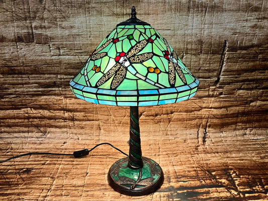 Tiffany-stijl tafellamp met een kleurrijke glazen kap in groen- en blauwtinten, versierd met gedetailleerde libellen met rode ogen; de lamp staat aan en verspreidt warm licht dat het mozaïekpatroon accentueert, met een decoratieve voet.