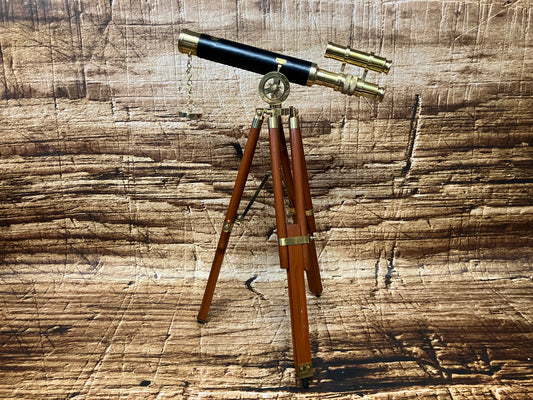 Een vintage telescoop met een zwarte buis en glanzende messing details staat op een elegante houten driepoot. Aan de zijkant zit een kleine zoekerscope, eveneens van messing. Een fijn kettinkje met een rond gewichtje hangt decoratief onder de hoofd­buis. De warme houttinten van het statief en de verweerde houten achtergrond versterken de klassieke, bijna maritieme uitstraling van het instrument.