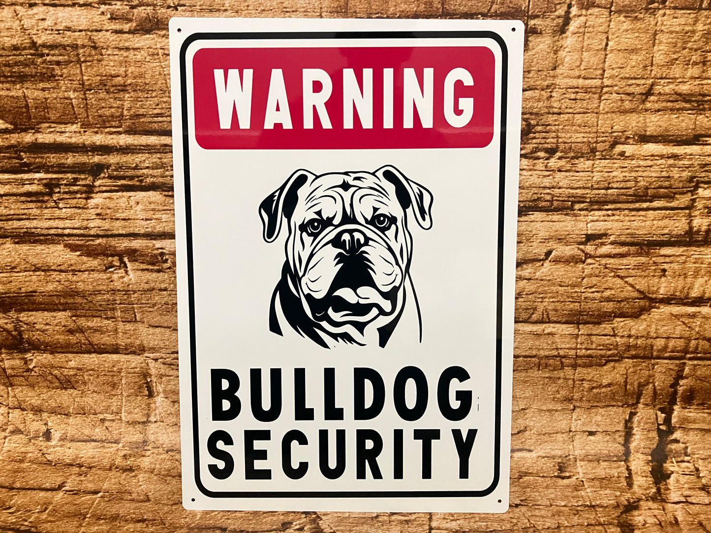 Waarschuwingsbord met een rode kopregel met het woord “Warning”, daaronder een zwart‑wit illustratie van een bulldog met een stoere, alerte blik. Onder de afbeelding staat in grote zwarte hoofdletters “Bulldog Security”. Het bord heeft een zwarte rand en hangt tegen een houten achtergrond, waardoor het geheel een robuuste, licht humoristische beveiligingsuitstraling krijgt.