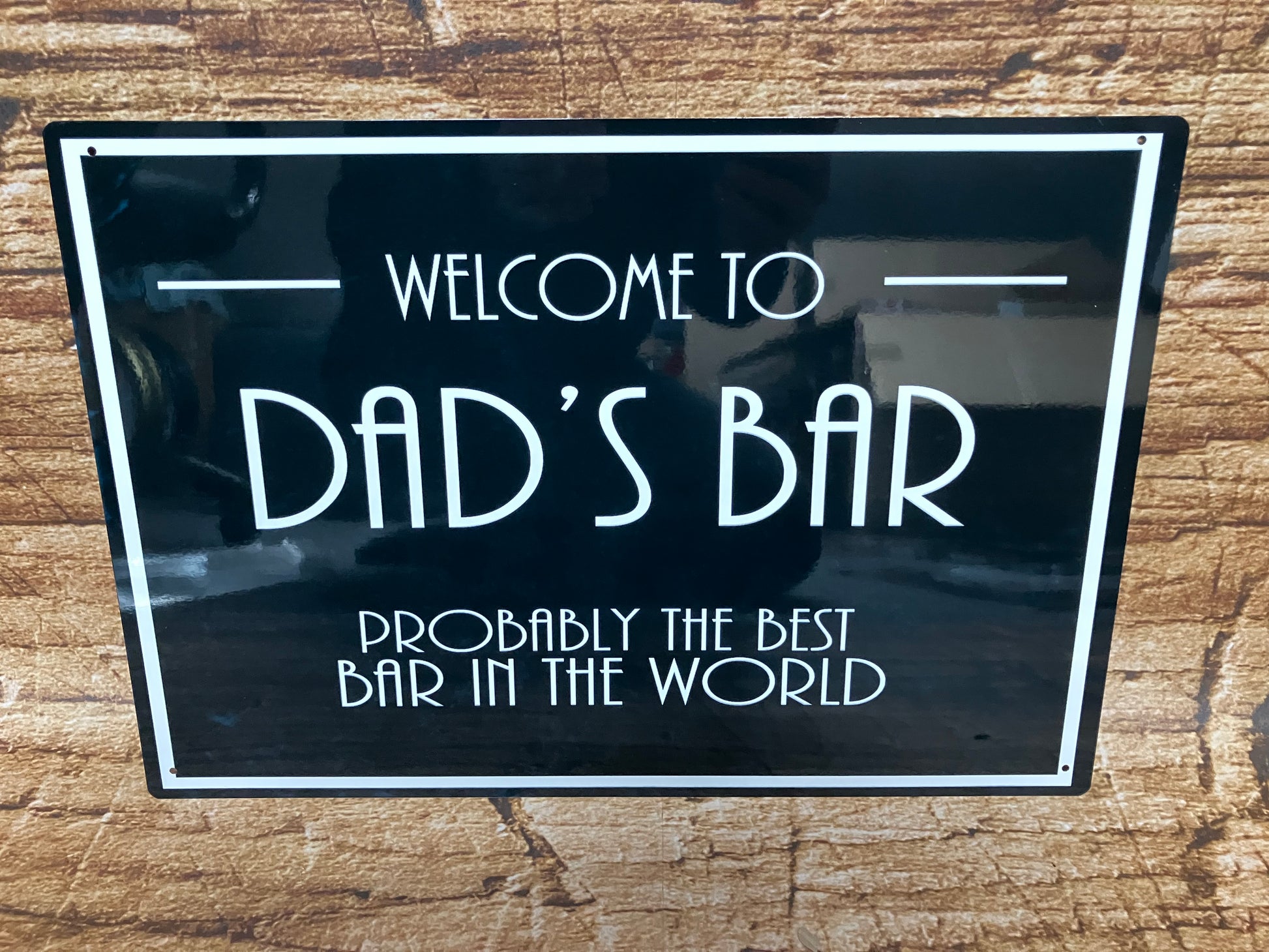 Decoratief bord met witte tekst “Welcome to Dad’s Bar – Probably the best bar in the world” op zwarte achtergrond met witte rand, gemonteerd op hout voor een gezellige en humoristische sfeer.